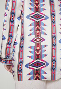 Blouse à motifs colorés présentant des motifs géométriques en rouge, bleu et noir sur un fond clair. Tissu lisse avec des accents contrastants.