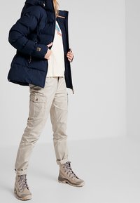 Tmavě modrá naducaná bunda, krémová košile s grafickým motivem, světle béžové kalhoty typu cargo a šedé turistické boty se šněrováním.