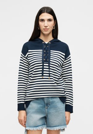 Donna con capelli lunghi e scuri che indossa orecchini a cerchio dorati, felpa a righe blu navy e bianca con dettagli con lacci e pantaloncini in denim azzurro chiaro sfrangiati.