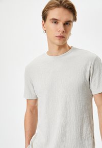 T-shirt corto a maniche corte di un grigio chiaro con texture, colletto rotondo e motivo sottile. Il tessuto appare morbido e leggermente sgualcito.