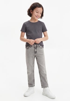 Kind mit kurzen dunklen Haaren, das ein dunkelgraues T-Shirt, hellgraue Jeans und weiße Sneakers trägt, steht und lächelt.