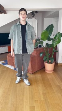 Camisa de mezclilla gris de botones sobre una camiseta negra, combinada con pantalones grises y zapatillas deportivas plateadas. Planta en maceta y un sofá al fondo.