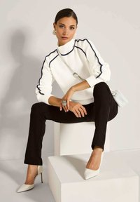 Frau in weißem Rollkragensweater mit blauen Streifen, schwarzen Hosen, weißen High Heels, großem Ring und weißer Umhängetasche, die auf einem weißen Block sitzt.