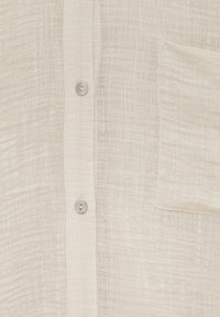 Tissu texturé beige clair avec patte de boutonnage verticale et deux petits boutons ronds, doté d'une poche plaquée rectangulaire du côté droit.