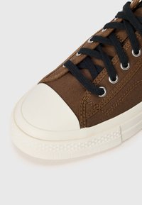 Bruine canvas sneaker met een witte rubberen neuskap, zwarte veters en metalen oogjes. Getextureerde details op de zool en zijkant.