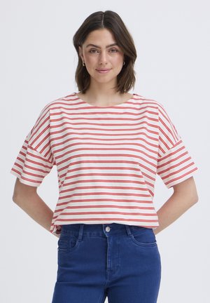 Mladá žena s krátkými hnědými vlasy, oblečená v bílém a červeně pruhovaném tričku s krátkými rukávy a tmavě modrých džínách, stojící před jednobarevným pozadím.