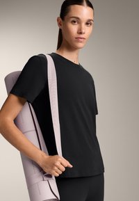 Mladá žena v černém cvičebním oblečení nese přes rameno stočenou růžovou jógamatku na neutrálním pozadí.