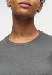 Κοντινό πλάνο του κατώτερου μέρους του προσώπου ενός ατόμου, του λαιμού και γκρι ριμπ πλεκτού πουκαμίσου, με εμφανείς πολλαπλές τρυπήσεις στα αυτιά και ασημένια σκουλαρίκια σε σχήμα κρίκου.