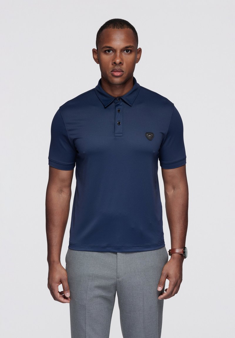 Granatowa koszulka polo z zaostrzonym kołnierzykiem, pliską na trzy guziki i małym czarnym logo na piersi. Materiał wydaje się gładki i dopasowany.