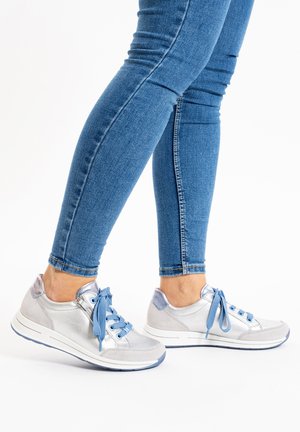 Gambe con jeans skinny blu e scarpe da ginnastica argento con lacci blu, in piedi su sfondo bianco.