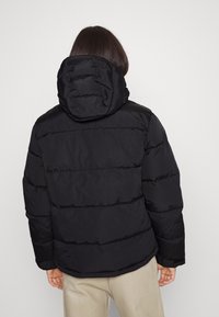 Giacca a piumino nera con cappuccio, design trapuntato e polsini. Il tessuto ha una superficie liscia con una vestibilità funzionale e rilassata.