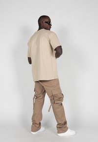 Beige bomulls t-skjorte og brune cargo-bukser med flere lommer. Modellen har på seg svarte solbriller og hvite sneakers, stående mot en lys bakgrunn.