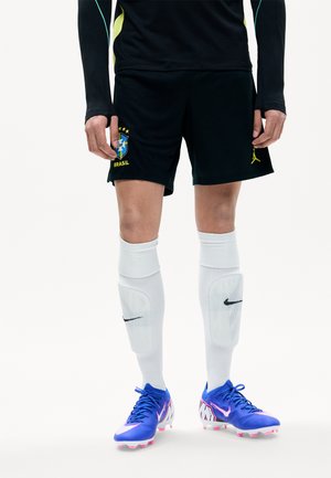 Persona che indossa pantaloncini da calcio del Brasile neri, parastinchi e calzini bianchi Nike, e scarpe da calcio Nike blu-rosa, in piedi su sfondo bianco.