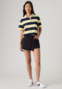 Νεαρή γυναίκα που στέκεται φορώντας κίτρινη και ναυτικό ριγέ πόλο μπλούζα, μαύρη φούστα τύπου κρουαζέ και λευκά αθλητικά παπούτσια μπροστά σε απλό φόντο.