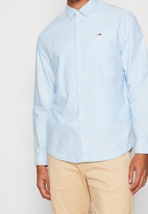 Homme portant une chemise bleu clair à manches longues avec un petit logo sur la poitrine et un pantalon beige, debout devant un fond uni.