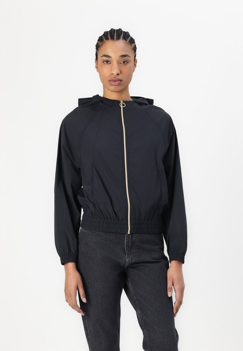 Giacca leggera nera con cappuccio, chiusura frontale con zip dorata, polsini elasticizzati e orlo arricciato. Texture liscia, design minimalista.
