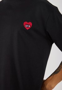 Black cotton T-shirt with a red embroidered heart featuring an eye in the center. The fabric is smooth with a classic crew neckline.