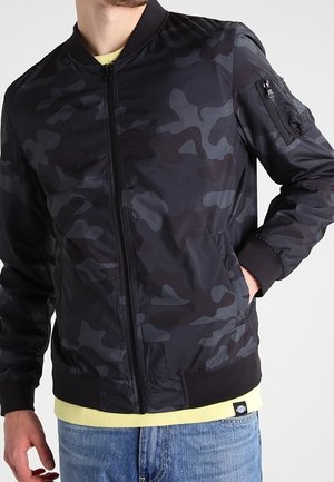 Mann trägt eine schwarze und graue Tarn-Bomberjacke mit Reißverschlusstasche am Ärmel, hellgelbes Hemd und blaue Jeans.