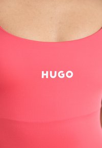 Ružový top bez rukávov vyrobený z hladkej tkaniny, s okrúhlym výstrihom a slovom "HUGO" v tučných bielych písmenách na hrudi.