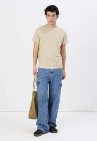 Pruhované béžové tričko a volné modré denimbordové kalhoty. Model drží olivově zelenou tašku a má na sobě černé boty. Jednoduchý pozadí.