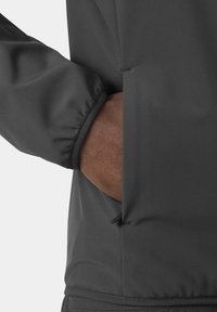 Mano de piel morena dentro del bolsillo de una chaqueta gris oscuro con puño elástico y detalle de cremallera.