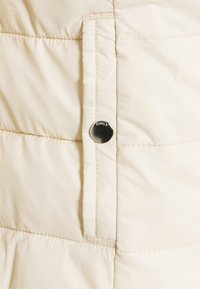 Zbliżenie beżowej tkaniny pikowanej z przyszytymi szwami oraz okrągłym srebrnym guzikiem z wygrawerowanym napisem "ONLY" na płaszczu lub kurtce.