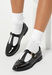 Czarne lakierowane buty na pasku typu T z perforowanymi zdobieniami, zestawione z białymi, koronkowymi skarpetkami do kostek, prezentujące eleganckie wykończenie.