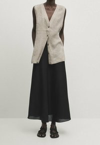 Beige ärmellose Leinenweste mit V-Ausschnitt, versehen mit drei strukturierten Knöpfen, kombiniert mit einem langen schwarzen Rock und flachen Sandalen.