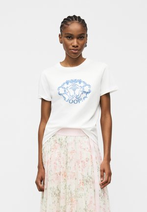 Sieviete ar pītu matu sakārtojumu, tērpusies baltā T-kreklā ar zilā emblēmu un ziedu pasteļtoņu svārkos, stāvot pret vienkāršu gaišu fonu.