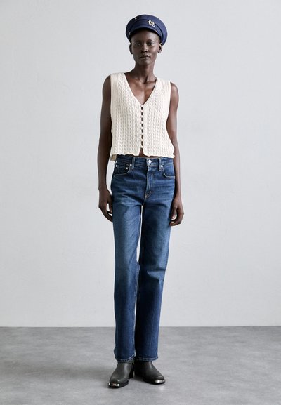 Crème kabelgebreide crop top, high-waisted donkerblauwe jeans en zwarte hakken. Model draagt een marineblauwe baret met gouden accenten.
