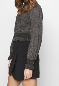 Kvinna som bär en metallgrå långärmad blus med volangade ärmslut och en svart hög midja kjol med en framficka.