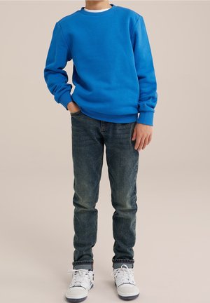 Lapsi, joka on pukeutunut siniseen pitkähihainen collegepaitaan, tummiin resoreilla varustettuihin farkkuihin ja valkoisiin tennareihin, seisoo yhdellä kädellä taskussa neutraalia taustaa vasten.