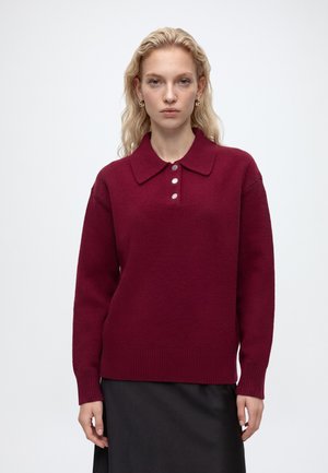 Vrouw met lang blond haar draagt een bordeauxrode gebreide trui met een brede kraag en drie knopen, gecombineerd met een zwarte rok.