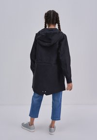 Zwarte waterdichte jas met een capuchon, elastische taille en lange mouwen, gecombineerd met blauwe jeans en zilveren sneakers. Zachte, gladde textuur.