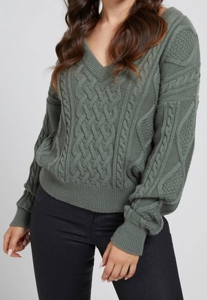 Grüner Strickpullover mit V-Ausschnitt, der über Zopf- und Strukturmuster verfügt, gerippte Bündchen hat und eine lockere Passform bietet. Kombiniert mit schwarzen Hosen.