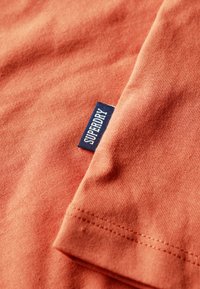 Lys orange bomuldsstof med en struktureret, ribbet overflade. Har et blåt "SUPERDRY" mærke syet på kanten.