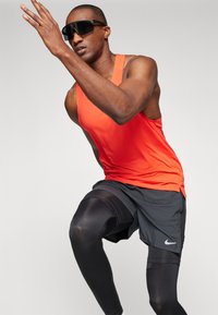 Conjunto deportivo masculino que incluye una brillante camiseta sin mangas naranja y pantalones cortos negros sobre leggings de compresión negros; incluye gafas de sol.