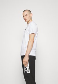 Camiseta blanca de algodón con mangas cortas, combinada con pantalones negros que tienen un gran logo blanco. El modelo está de perfil con una pose neutral.
