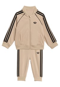 Bež trenirka s črnim dizajnom s tremi črtami, jakna z zadrgo in rebrastim ovratnikom ter ujemajoče hlače z rebrastimi manšetami in logotipom.