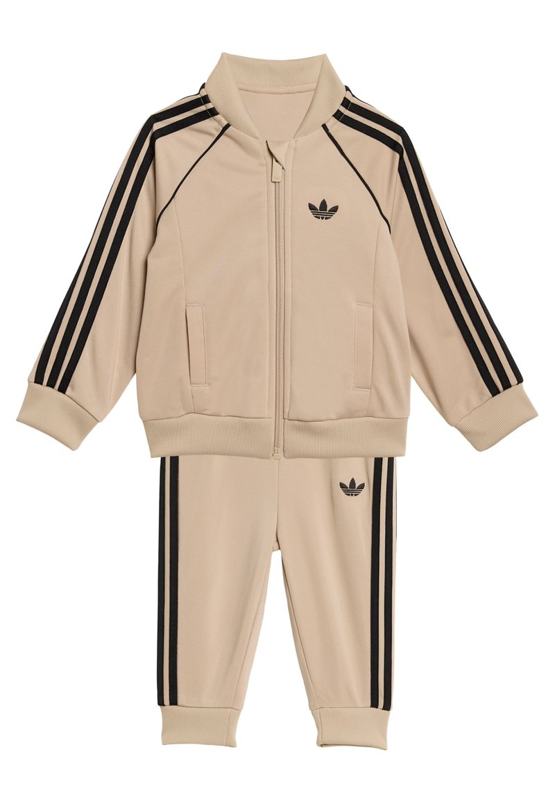 Bež trenirka s črnim dizajnom s tremi črtami, jakna z zadrgo in rebrastim ovratnikom ter ujemajoče hlače z rebrastimi manšetami in logotipom.