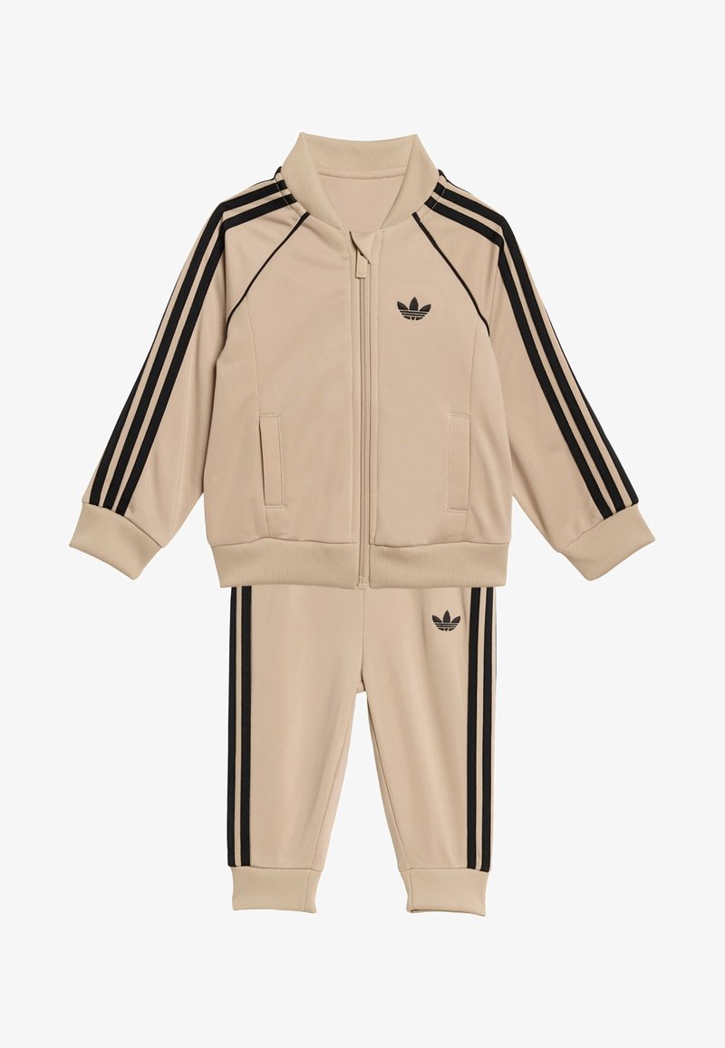 Bež trenirka s črnim dizajnom s tremi črtami, jakna z zadrgo in rebrastim ovratnikom ter ujemajoče hlače z rebrastimi manšetami in logotipom.