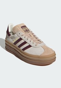 Tenisky Adidas Gazelle s béžovým a vínovým semišovým zvrškom, dizajnom troch pruhov a gumovou podrážkou s embosovaným brandingom.