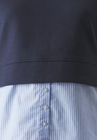 Haut bleu marine superposé à une chemise à boutons bleu clair à rayures verticales, avec trois boutons visibles. Tissu en coton, design décontracté.