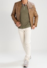 Chaqueta de cuero marrón con detalles texturizados, camiseta henley verde, chinos de color crema claro y zapatillas grises con acentos blancos.