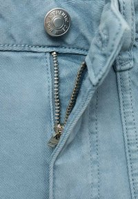 Svetlo modri kavbojke z vidnim kovinskim zadrgo delno odprto, opremljene z označenim gumbom in podrobnostmi teksturirane tkanine.