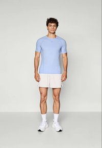 Junger Mann steht vor einem schlichten Hintergrund und trägt ein hellblaues Sportshirt, beige Shorts, weiße Socken und weiße Sneakers.