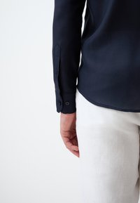 Marineblauw overhemd met lange mouwen en knoopmanchetten, gladde textuur en een getailleerde pasvorm, gecombineerd met witte linnen broek, gedeeltelijk zichtbaar.