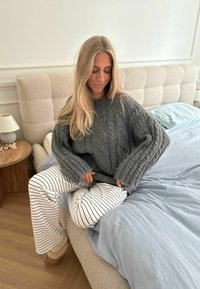 Maglione grigio lavorato a maglia con maniche a coste, abbinato a pantaloni bianchi a righe. Ambientazione della camera da letto con texture morbide e colori neutri.