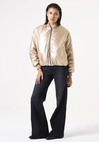 Veste bomber beige métallique avec une fermeture éclair à l'avant, des poignets et un ourlet côtelés. Associée à un jean noir à jambes larges et des chaussures noires.