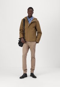 Giacca impermeabile marrone con cappuccio sopra una maglietta a righe, pantaloni beige e stivali neri. Tiene una borsa nera, in piedi davanti a uno sfondo semplice.